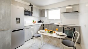 Een moderne keuken met een inbouwkoelkast, een magnetron en een fornuis. Er staat een eettafel met stoelen en erboven een raam.