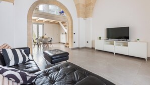 Un salon moderne avec un canapé en cuir, un téléviseur à écran plat posé sur un meuble blanc, et un coin repas à l’arrière-plan.