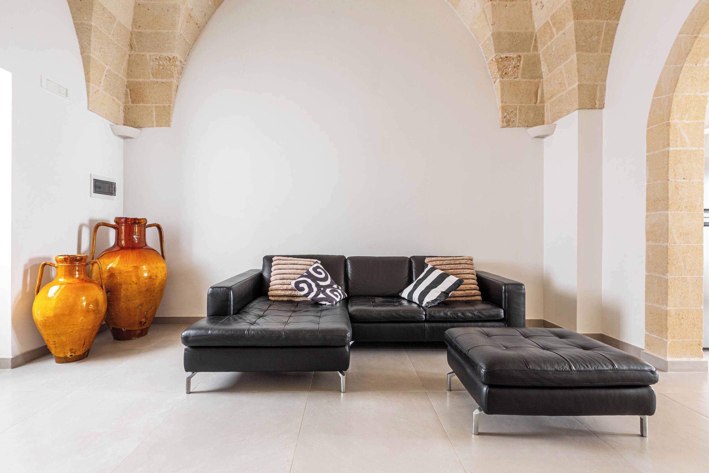 Ein modernes Wohnzimmer mit einem schwarzen Ledersofa, zwei passenden Hocker und zwei großen Vasen auf einem Fliesenboden.