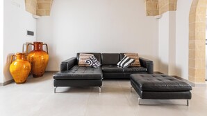 Un salon moderne avec un canapé en cuir noir, deux poufs assortis et deux grands vases posés sur un sol carrelé.