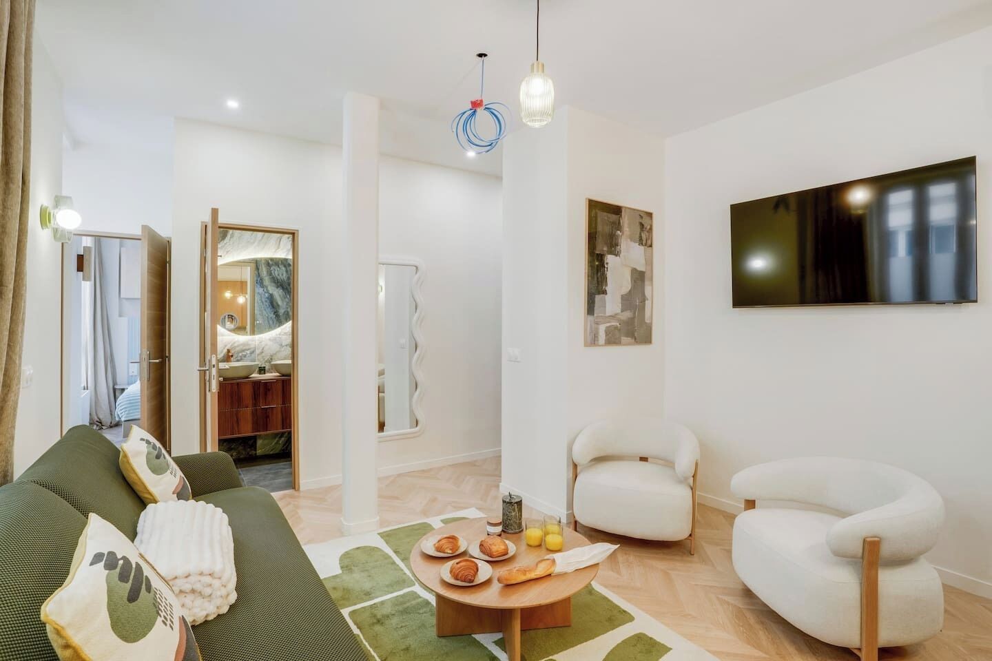 A modern living room with a sofa, chairs, a coffee table with pastries, and a TV on the wall.