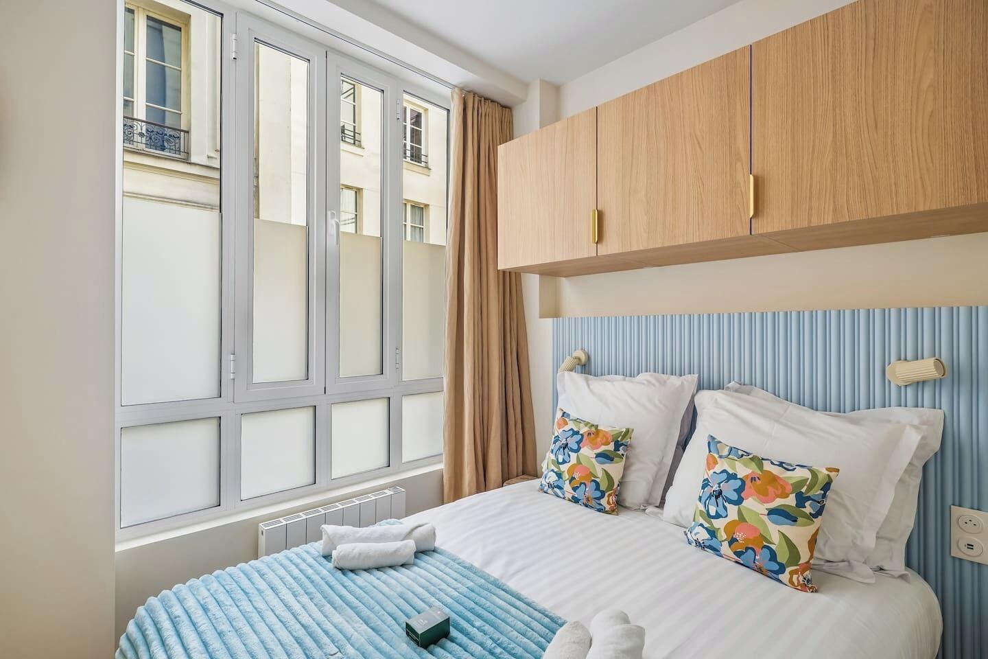 A hotel room with a large window, a bed with floral pillows, a blue-striped bedspread, and wooden headboard cabinets.