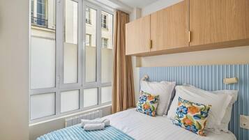 A hotel room with a large window, a bed with floral pillows, a blue-striped bedspread, and wooden headboard cabinets.