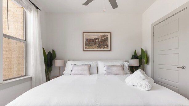 A bedroom with a large bed, two bedside lamps, a framed picture on the wall, and a window with curtains. - Comfortable 1BR Retreat at The Nelson TNS 5205 (Austin)