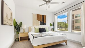 Ein Schlafzimmer mit Bett, Nachttischen, Deckenventilator, großem Fenster und Blick ins Freie.