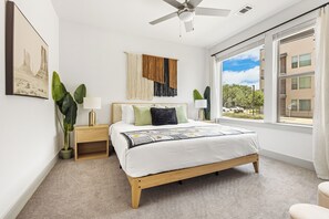 A bedroom with a bed, bedside tables, a ceiling fan, a large window, and a view of the outside. - Modern 2BR Haven with Balcony & Open Living (Austin)