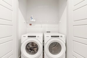 A laundry room with two front-loading Whirlpool washing machines and a shelf above them holding a bottle of Purex detergent. - Modern 2BR Haven with Balcony & Open Living (Austin)
