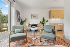 Ein modernes Wohnzimmer mit zwei Sesseln, einem runden Couchtisch und einem Esstisch mit Stühlen. Es gibt eine Küchenzeile mit Holzschränken und einer Spüle.