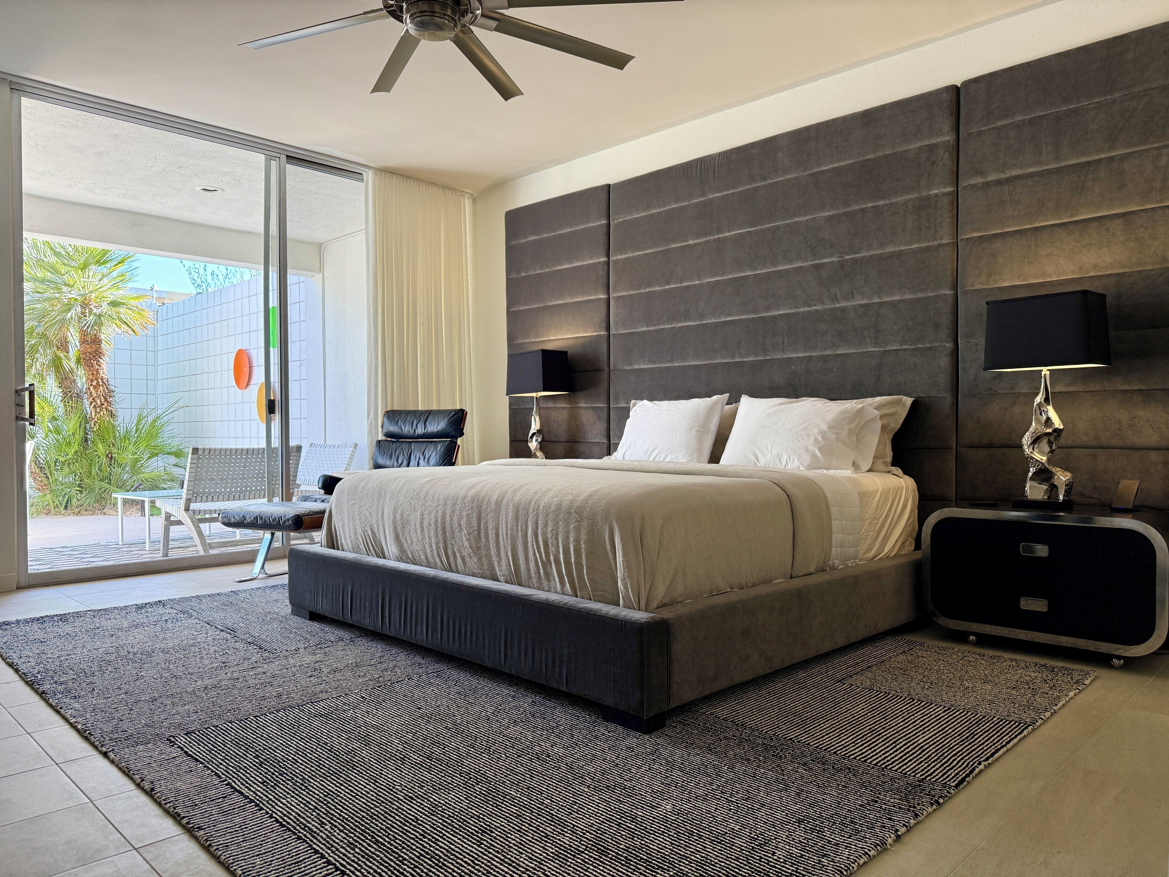 A modern bedroom with a large bed, a ceiling fan, a nightstand, and a view of the outdoors.