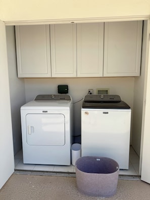 A laundry room with two white washing machines, a small trash bin, and a purple laundry basket. - Upscale 2-Bedroom + Den Condo in the Beautiful Seven Lakes Golf & Country Club (Palm Springs)
