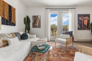 Een moderne woonkamer met een bank, fauteuil en een glazen salontafel. Er zijn twee ramen met gordijnen, een schilderij van een paard en een grote plant.
