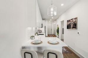 A modern kitchen with white cabinets, a dining area with a white table set for two, and a hallway leading to a door. - Studio-Style 1BR Retreat at The Nelson (Austin)