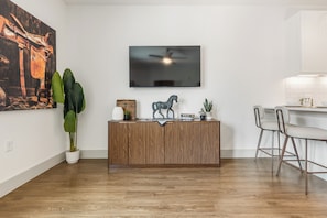 A modern living room with a wooden cabinet, a mounted TV, a dining area with a chair, and a large plant. - Studio-Style 1BR Retreat at The Nelson (Austin)