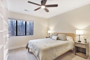 A bedroom with a bed, two bedside tables, a window with a view of trees, and a ceiling fan.