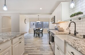 A modern kitchen with white cabinetry, granite countertops, and a dining area with a view of trees through the window.