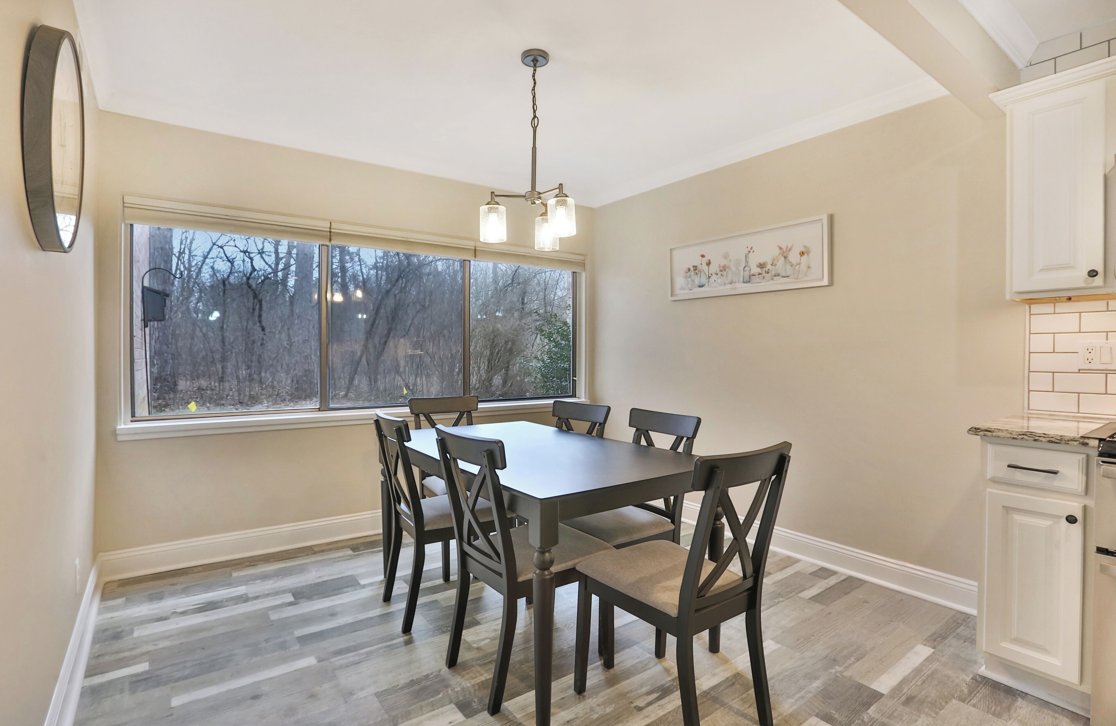 A dining room with a large table and chairs, a window with a view of trees, and a kitchen area with white cabinets.