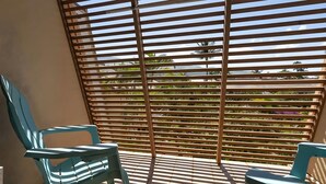A sunlit balcony with wooden slats, two turquoise chairs, and a view of greenery and buildings. - Ocean View 4BR, 2 Condos for 12 in Kihei! (Kihei)