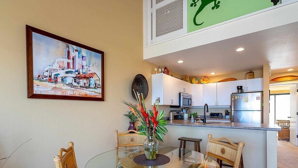 A dining area with a glass table, wicker chairs, and a painting on the wall. - Ocean View 4BR, 2 Condos for 12 in Kihei! (Kihei)