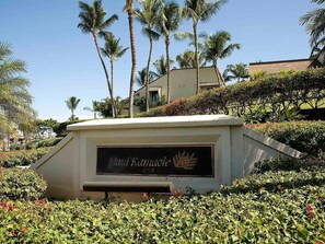 A landscaped area with a sign reading 'Min Kanoaole 2777' surrounded by greenery and palm trees. - Kihei, Maui Kamaole, Spacious 4 BR, Ocean View, Sleeps 12 (Kihei)