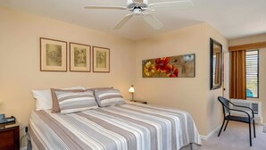 A bedroom with a bed, a desk, a chair, a ceiling fan, and artwork on the walls. - Ocean View 4BR, 2 Condos for 12 in Kihei! (Kihei)