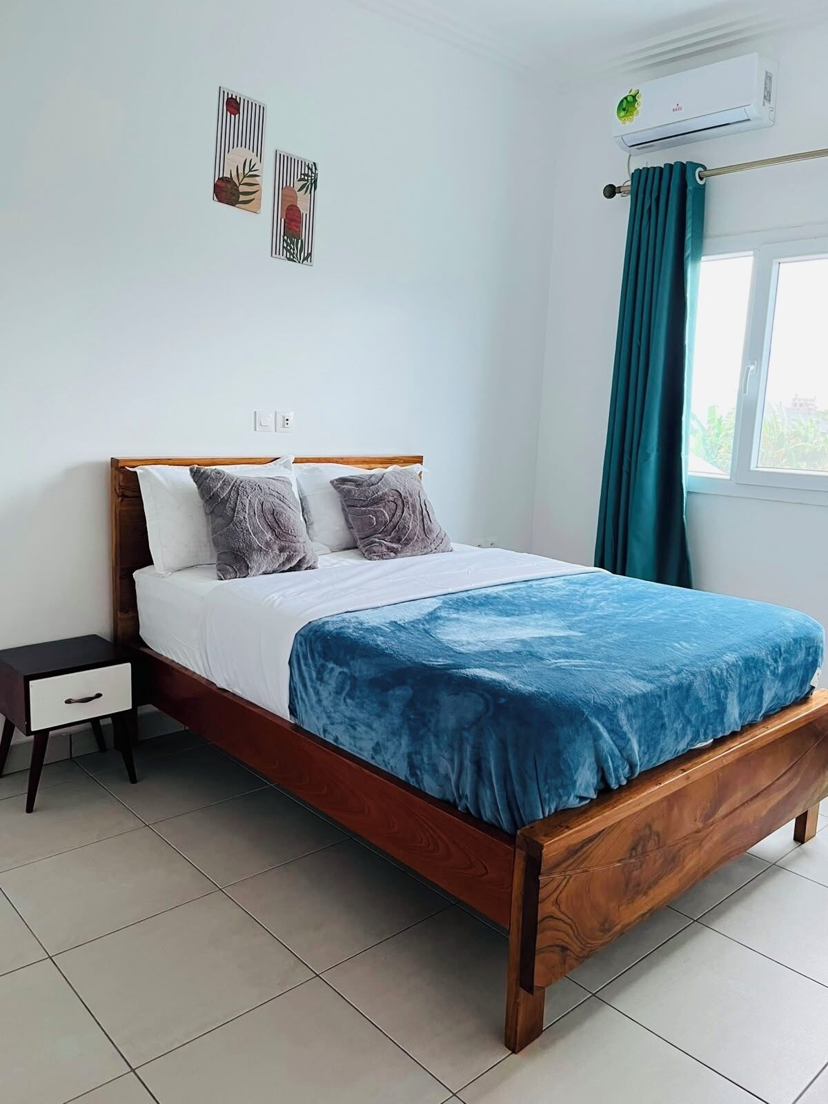 A single bed with a wooden frame, white bedding, and a blue blanket. There is a nightstand with a drawer, a wall-mounted air conditioner, and a window with curtains.