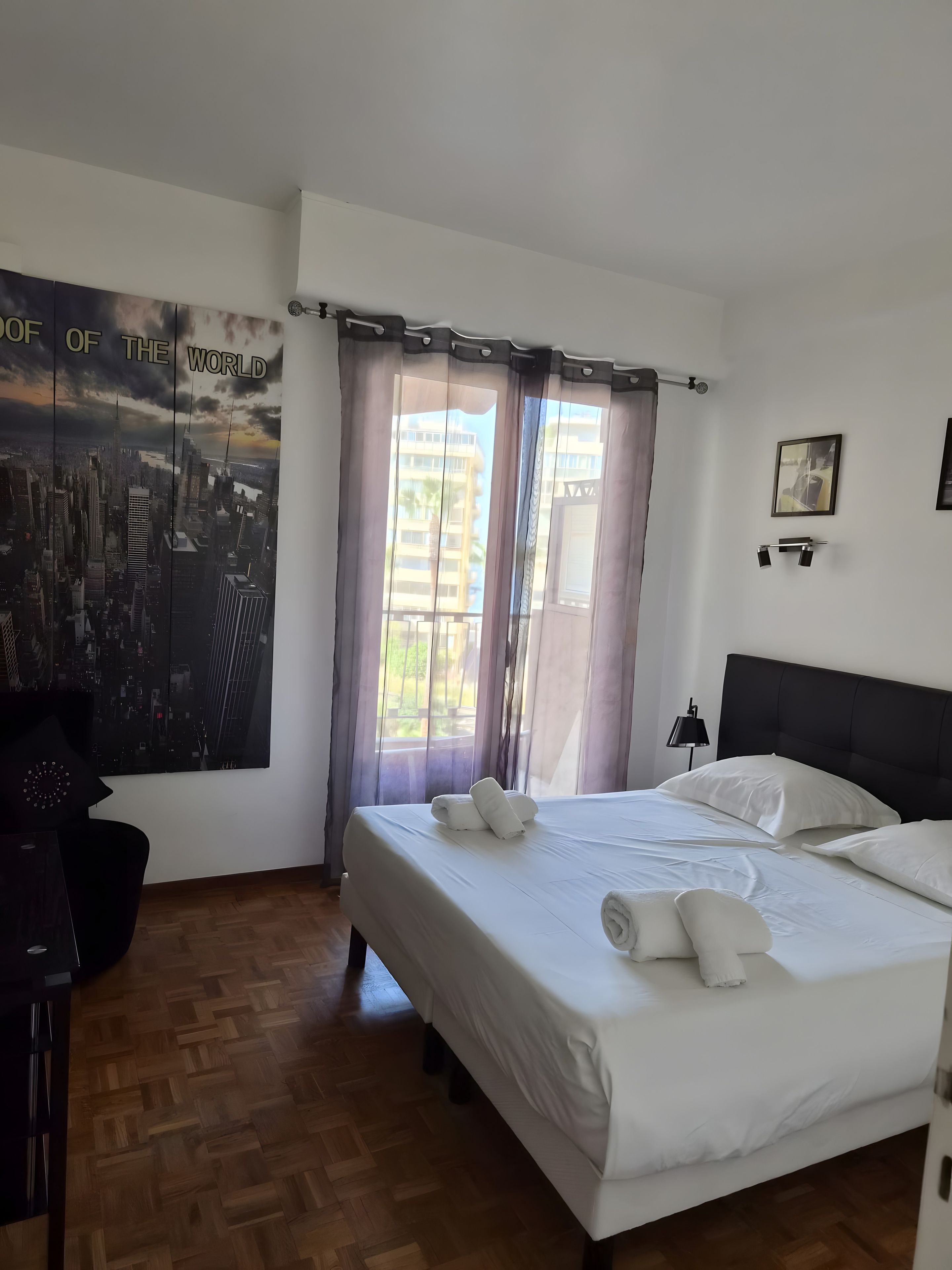 Ein Hotelzimmer mit einem großen Bett, Blick auf die Stadt durch transparente Vorhänge, Holzboden und einem schwarzen Kopfteil.