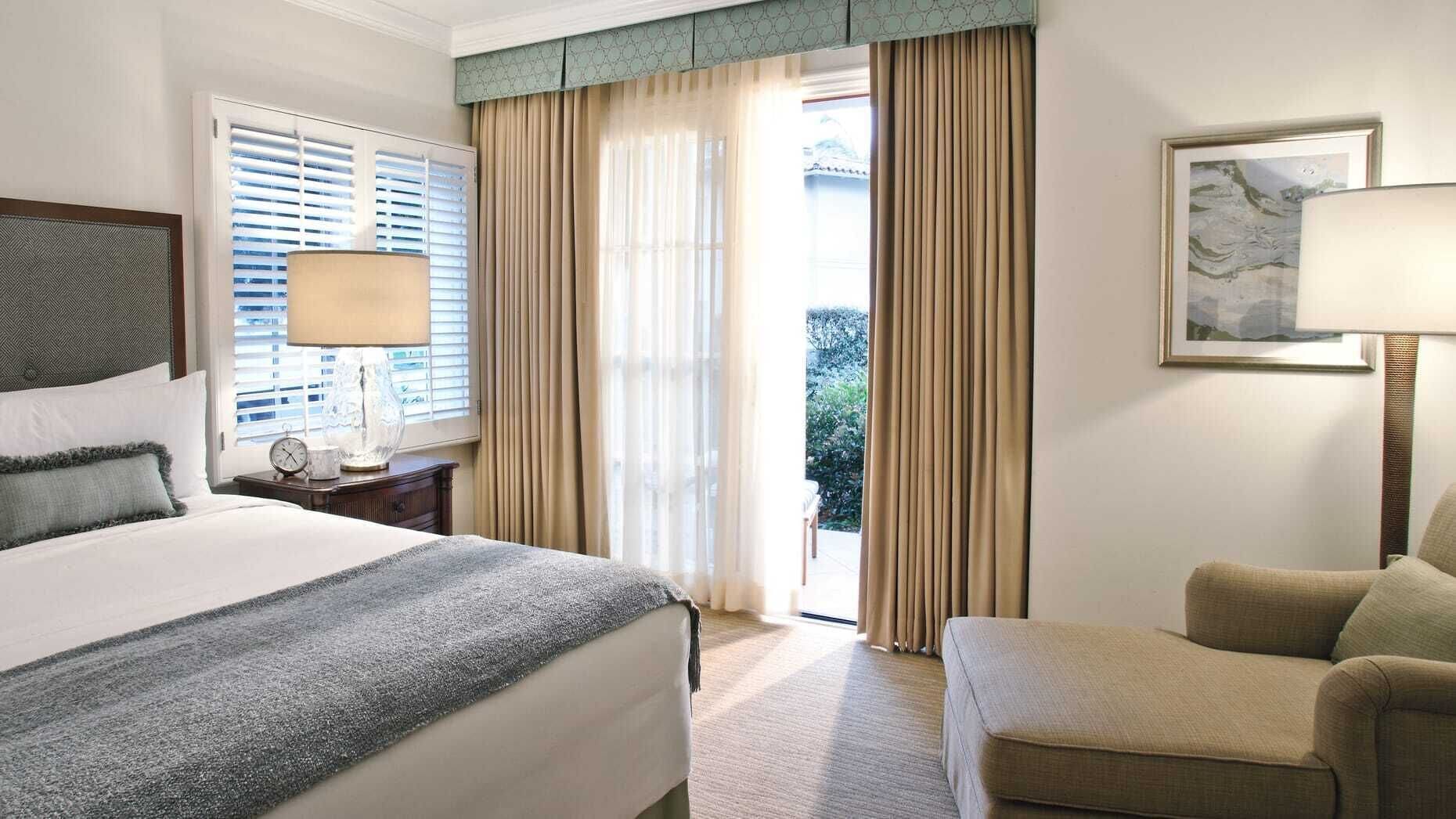 Habitación de hotel con cama, una silla, escritorio con lámpara, ventana con persianas y puerta que da a un balcón.