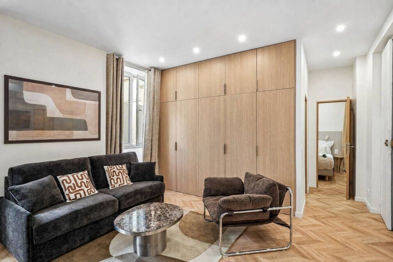 A modern living room with a sofa, armchair, and coffee table, a large wardrobe, and a painting on the wall.