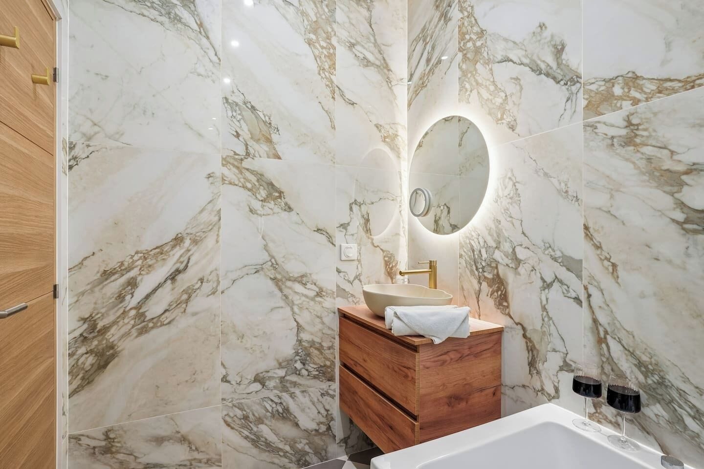 A bathroom with marble walls, a wooden vanity, a sink, a mirror, and a bathtub.