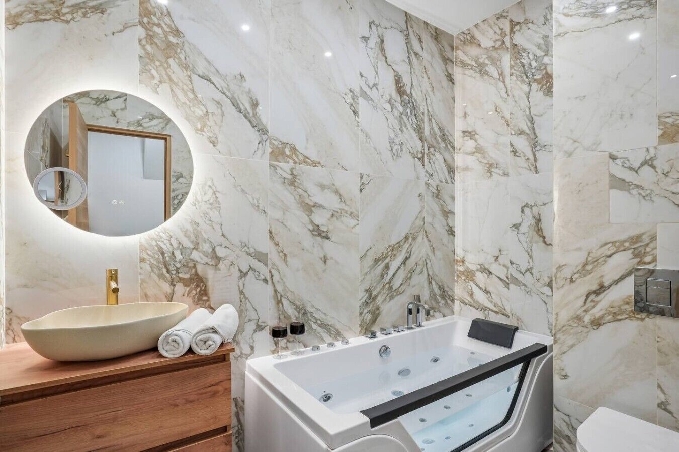 A bathroom with a marble wall, a round mirror, a sink, a bathtub, and a toilet.