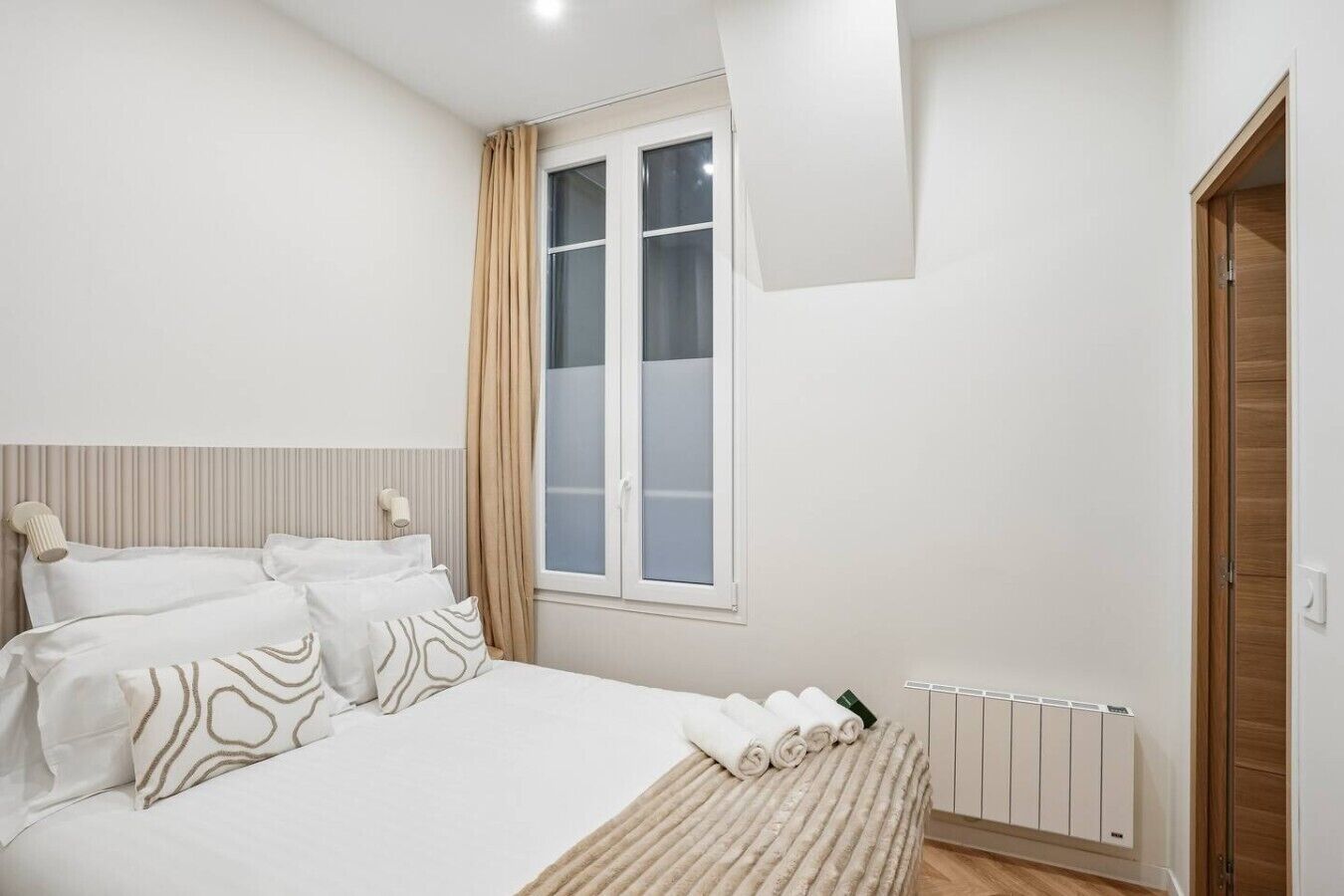 A bedroom with a bed, a window with curtains, a radiator, and a wooden door.