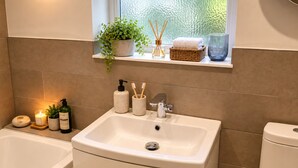 Un bagno con un lavandino bianco, una vasca da bagno e un water. Sopra il lavandino c'è uno scaffale con una pianta, una candela e un bicchiere.