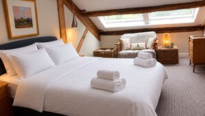 Una camera d'albergo con un letto grande, un comodino, una poltrona in vimini, un tavolino con una lampada e un settimino in legno.