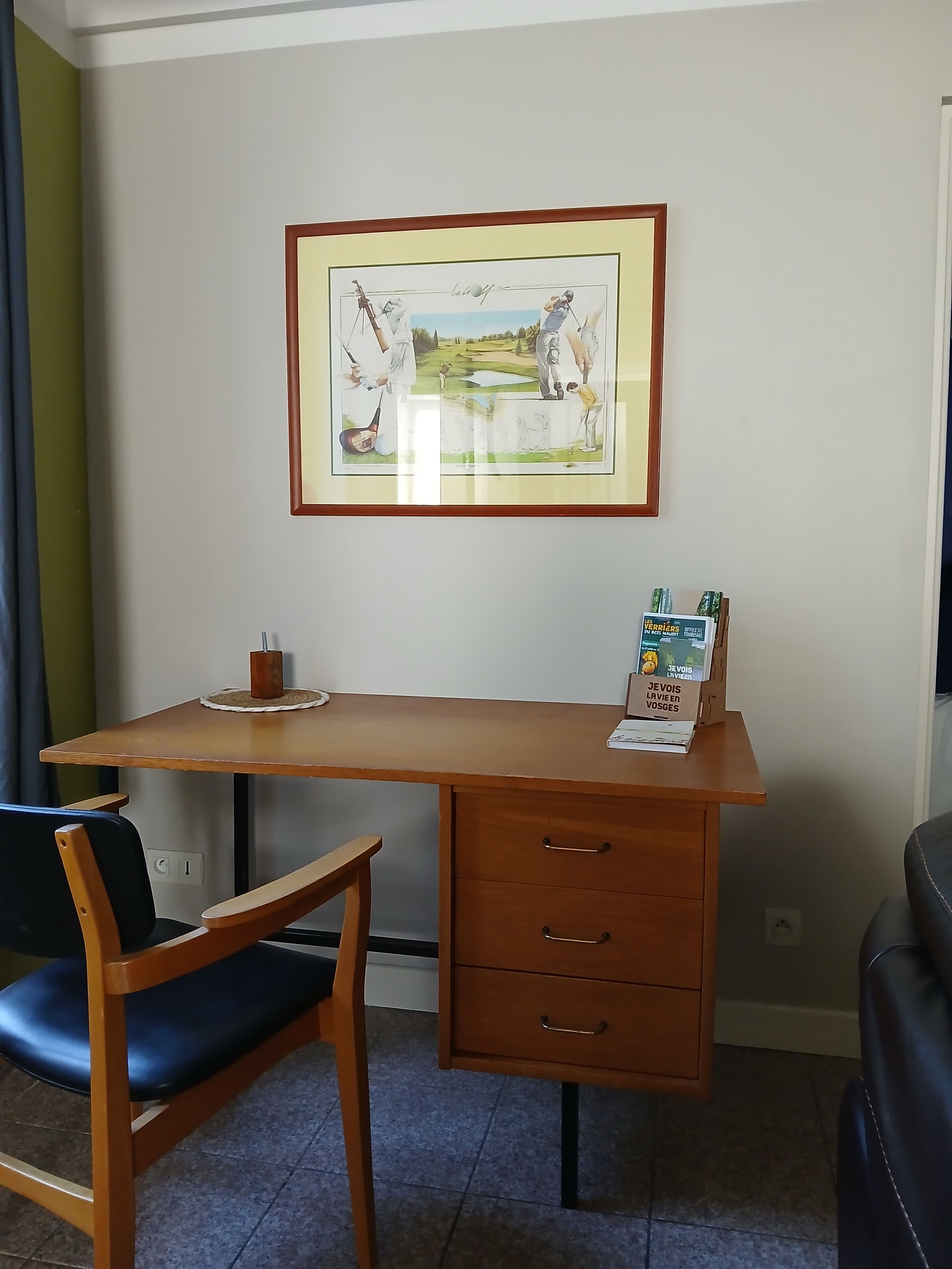 A room with a wooden desk, a chair, a framed picture on the wall, and a small table with a box on it.