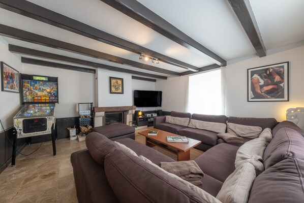 A living room with a large sectional sofa, a fireplace, a flat-screen TV, and a pinball machine.