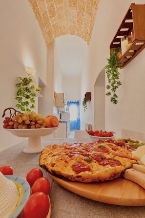 Ein Essbereich mit einer großen Pizza auf einem Holzbbrett, einer Obstschale und einer Pflanze auf einem Regal.