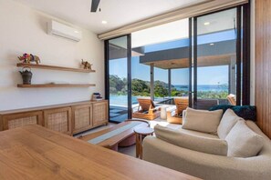 A modern living room with a wooden table, sofa, and chairs, large sliding glass doors leading to a balcony with a view of the sea. - Oceanview Luxury Home in Pacifico Coastal Living (Coco)