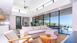 A modern living room with a large sectional sofa, wooden chairs, a coffee table, and a view of the ocean through floor-to-ceiling windows. - Oceanview Luxury Home in Pacifico Coastal Living (Coco)