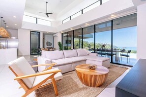 A modern living room with a large sectional sofa, wooden chairs, a coffee table, and a view of the ocean through floor-to-ceiling windows. - Oceanview Luxury Home in Pacifico Coastal Living (Coco)