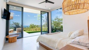 A modern bedroom with a large bed, a wooden side table, a TV mounted on the wall, and a large sliding glass door leading to an outdoor area with a view of the sea. - Oceanview Luxury Home in Pacifico Coastal Living (Coco)