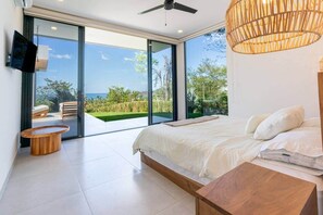 A modern bedroom with a large bed, a wooden side table, a TV mounted on the wall, and a large sliding glass door leading to an outdoor area with a view of the sea. - Oceanview Luxury Home in Pacifico Coastal Living (Coco)
