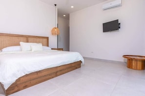 A bedroom with a wooden bed, white bedding, a hanging light fixture, a television on the wall, and a small wooden table. - Oceanview Luxury Home in Pacifico Coastal Living (Coco)