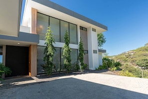 A modern two-story house with large glass windows, a paved driveway, and a landscaped garden with tall trees. - Oceanview Luxury Home in Pacifico Coastal Living (Coco)