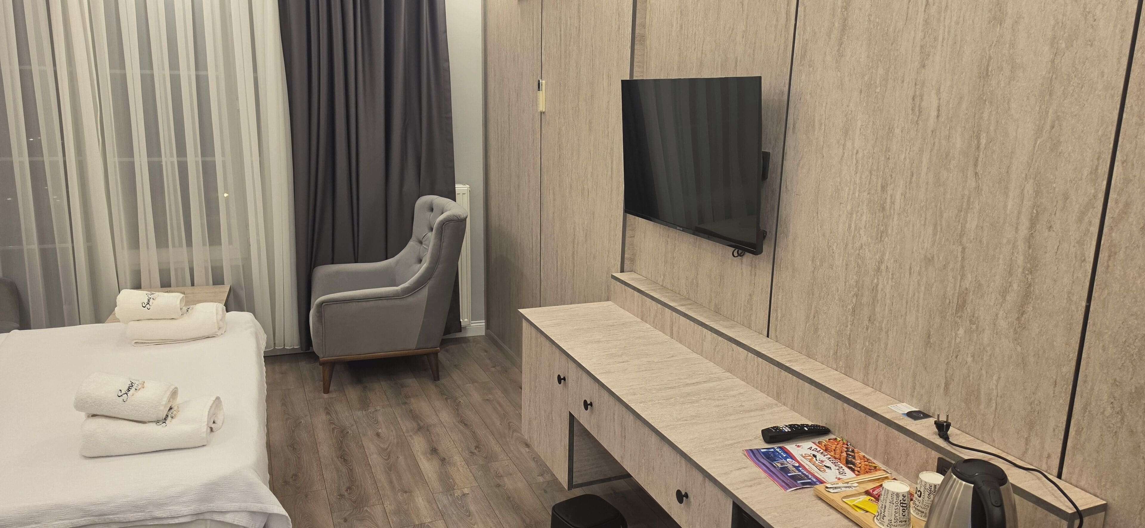 A hotel room with a bed, a grey armchair, a TV mounted on the wall, a wooden desk, and a coffee machine.