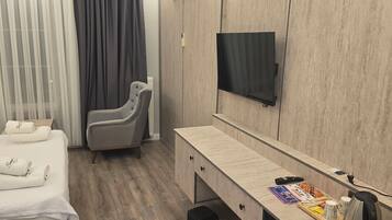 A hotel room with a bed, a grey armchair, a TV mounted on the wall, a wooden desk, and a coffee machine.