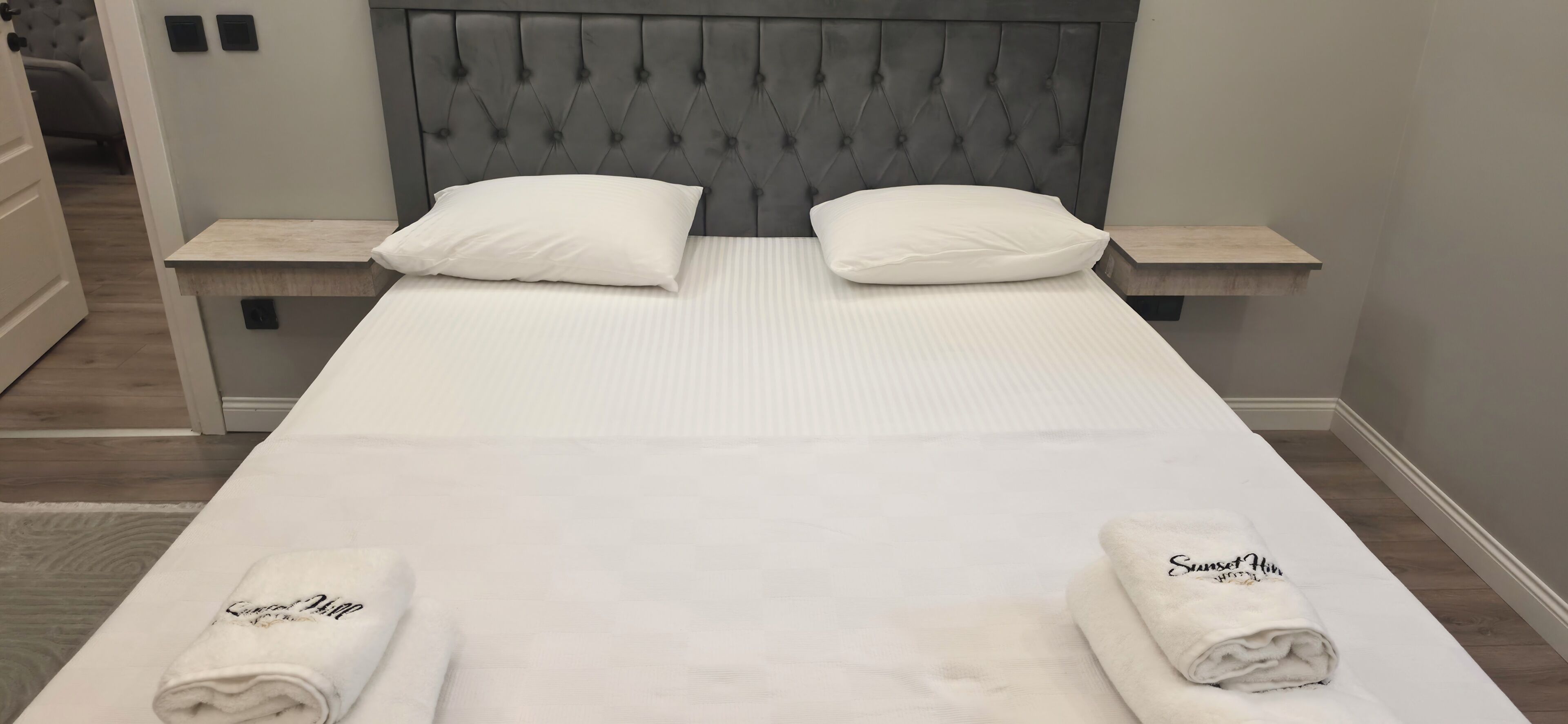 A double bed with a tufted headboard, two bedside tables, and two rolled towels with text on them.