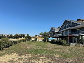 Un barrio residencial con una amplia casa de dos pisos, balcón, césped y cielo azul despejado.
