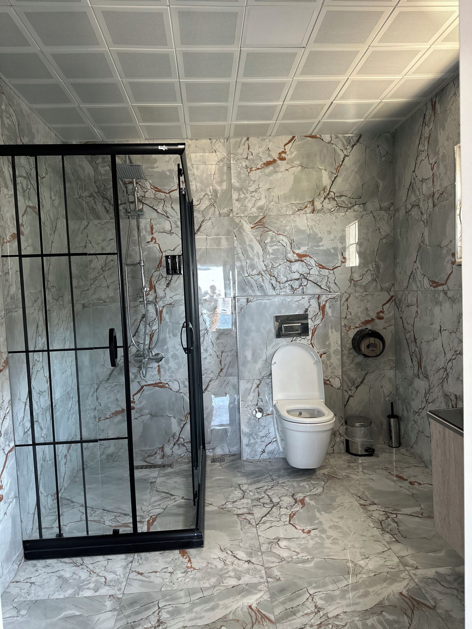 A bathroom with a marble floor, a toilet, a shower enclosure, and a ceiling with a grid pattern.