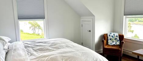 A bedroom with a large bed, a chair, and two windows with blinds.