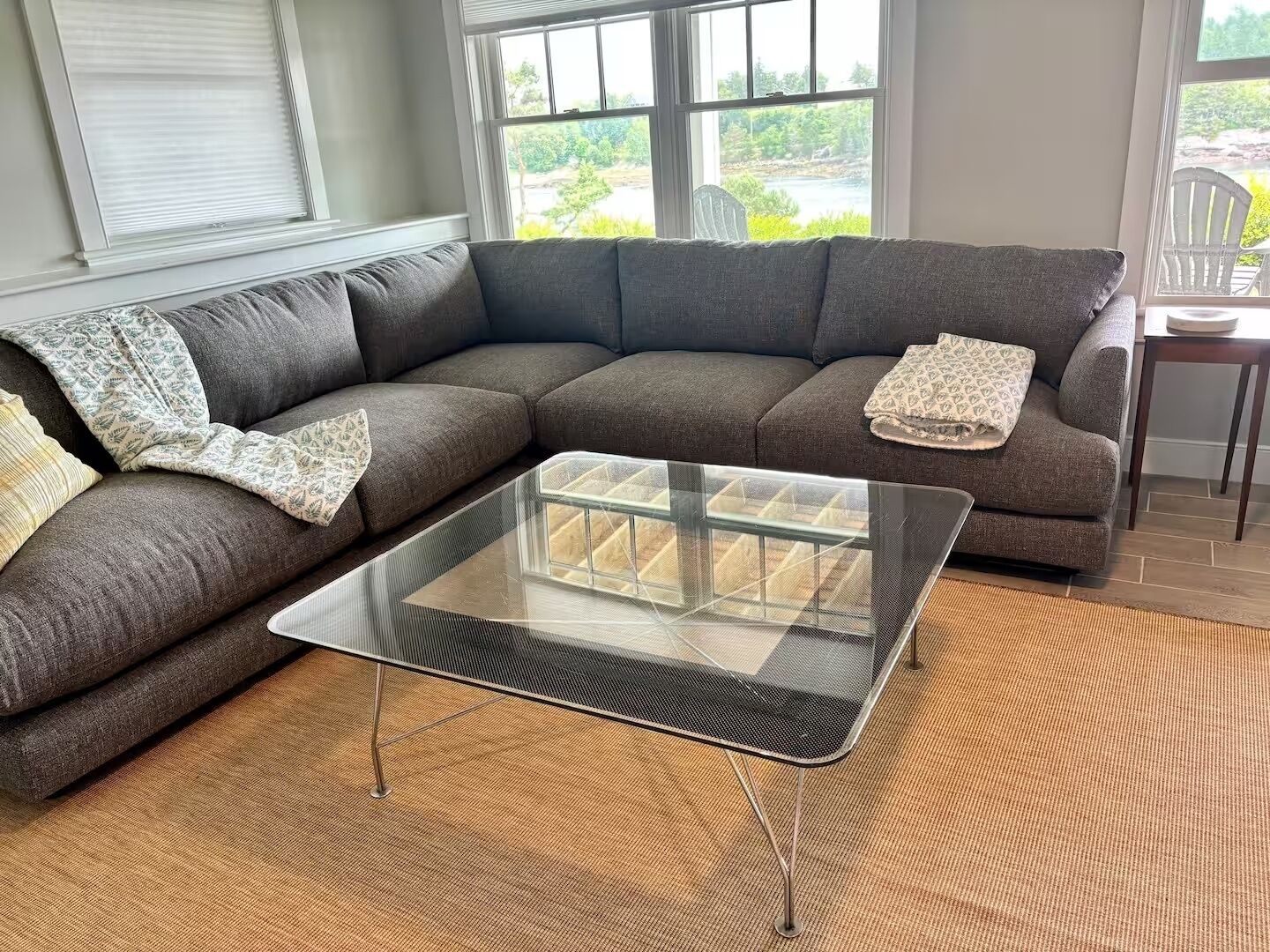 A living room with a sectional sofa, a glass coffee table, and a view through large windows.